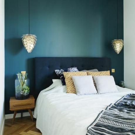 Stilvoller Schlafzimmerinnenraum mit kleinem hölzernem Nachttisch, Garten in einem Glas, weißer Bettwäsche, Farbpilows und Decke. Raum mit blauen Wänden und braunem hölzernem Parkett. Design Lampe.
