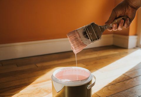 Hand hält einen dicken Pinsel und entfernt ihn, nach dem Eintauchen in die Farbe entsteht ein Farbtropfen, der spritzt zurück in den Topf die Wand dahinter ist orange gestrichen und die Farbe in der Dose ist pink, was konzeptionell mit Platz für kollidiert Kopieren