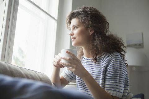 Lächelnde junge Frau mit dem Tasse Kaffee, der aus Fenster heraus schaut