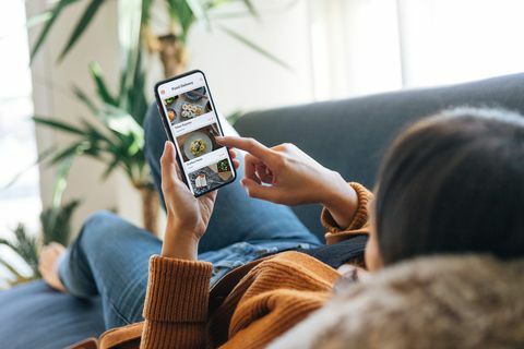 über die Schulteransicht einer jungen Frau, die Essen von der Speisekarte auf der mobilen App auswählt, während sie zu Hause auf der Couch liegt