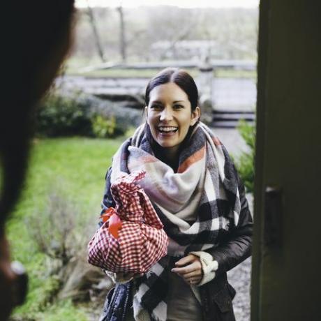 eine Frau lächelt, als sie auf der Türschwelle steht und eine Freundin besucht sie ist in warme Kleidung gehüllt und hält ein Geschenk in ein rot-weiß kariertes Tuch gehüllt