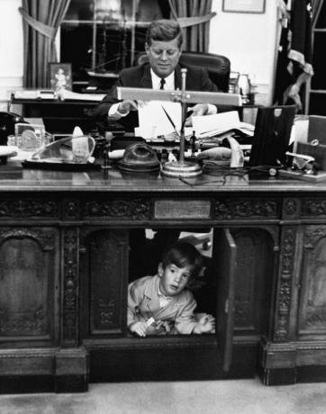 John F. Kennedy bei der Arbeit am entschlossenen Schreibtisch im ovalen Büro des Weißen Hauses, während sein Sohn, John F. Kennedy Jr., sich in der Einrichtung versteckt