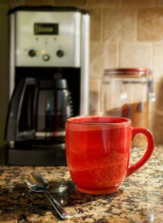 Dampfende Tasse Kaffee: Eine rote Tasse dampfenden Kaffees steht auf einer Küchenarbeitsplatte aus Granit. Eine Kaffeemaschine und ein Behälter mit gemahlenem Kaffee sind im Hintergrund zu sehen.