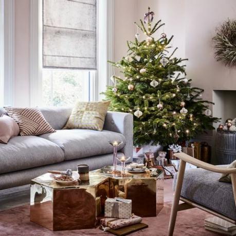 Weihnachtsbaum in der Ecke eines Wohnzimmers graues Sofa und goldene Sechseck-Couchtische, hellrosa Wände und graues Römer blind reflektierte Herrlichkeit atemberaubende Spiegel Messing Tische, Bronzeandpinkbaublesandaccentsofgoldbringamodernopulence toarelaxedlivingroom